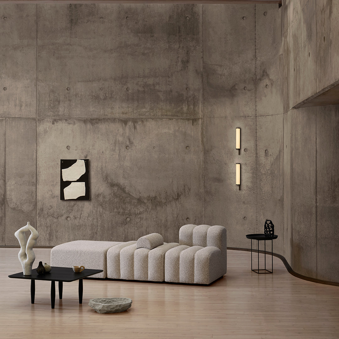 Minimalist beige sofa with black accents, set against a textured gray concrete wall, featuring clean lines and modern shapes.
