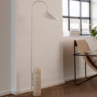 Minimalist floor lamp with a white shade and beige textured base, set against a light-toned room with wooden flooring.