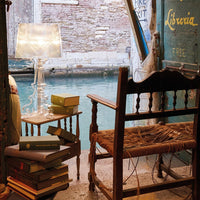 "Cozy scene with a rustic wooden chair, clear lamp, and vibrant books against a serene water backdrop."