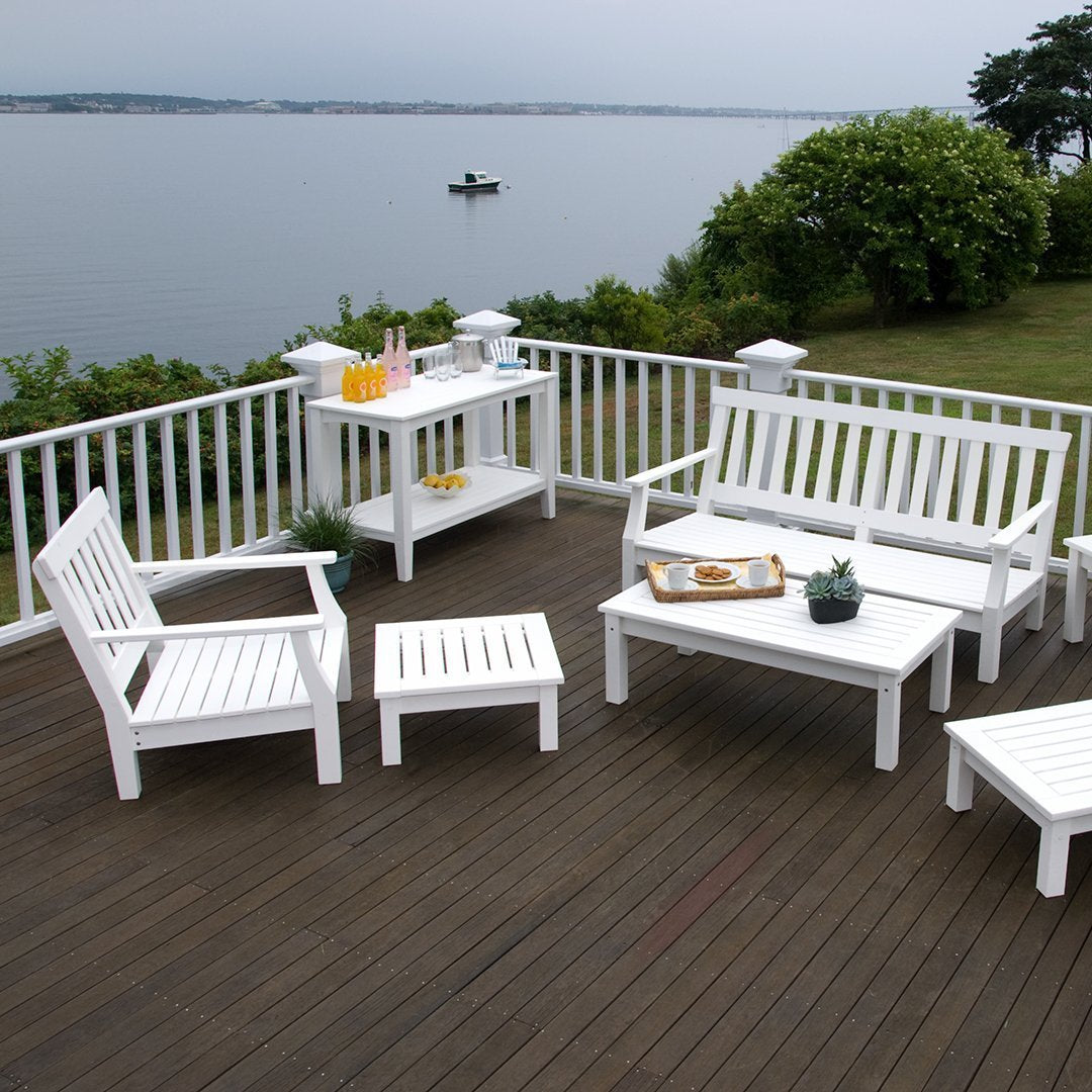 White patio furniture set on a natural wood deck, surrounded by green foliage and a serene water view.