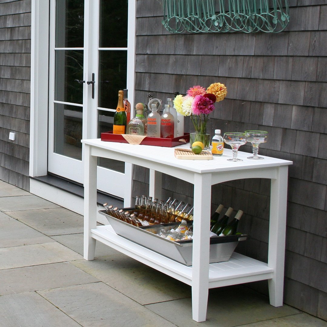 White outdoor serving table with colorful decor, including clear glass bottles, vibrant flowers, and drinks.