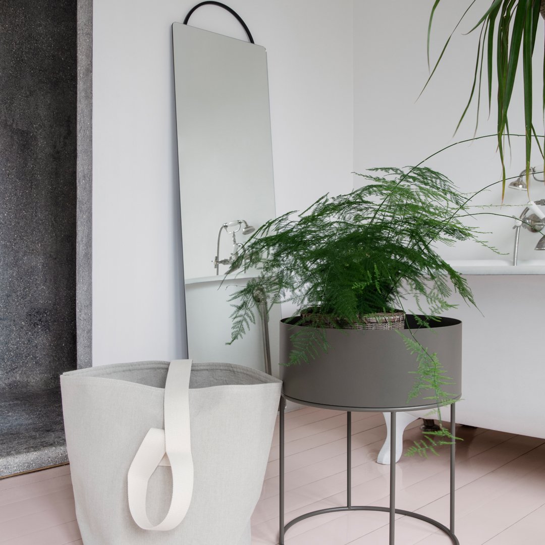 Light beige tote bag with a white handle, paired with a round gray planter and lush green foliage against a soft pink floor.