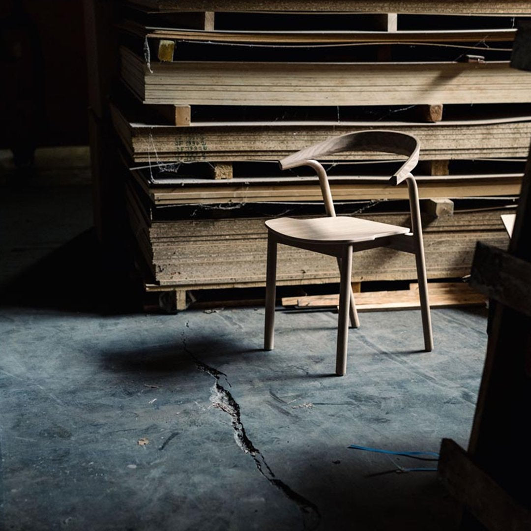 "Minimalist wooden chair in natural finish, set against a backdrop of stacked wood and dark floor."