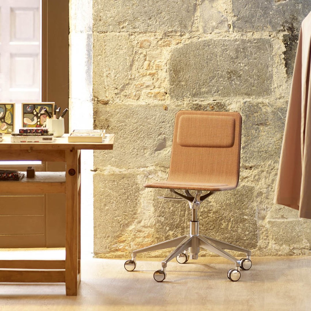 Modern swivel chair in warm tan fabric with a subtle textured pattern, set against a rustic stone wall backdrop.