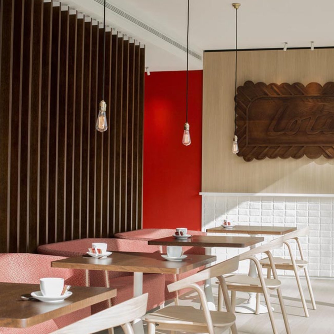 Modern cafe interior with red accent wall, wooden paneling, and light-colored furniture featuring subtle geometric patterns.