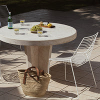 Round stone table with white wire chairs, set against beige tiles, featuring a natural woven basket and fresh fruit.