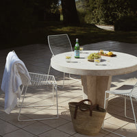 "White textured dining set with a round stone table and linear chairs, set against a natural outdoor backdrop."
