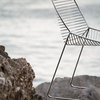 Sleek metal chair with a minimalist chrome finish, featuring string-like seat patterns against a natural rocky background.