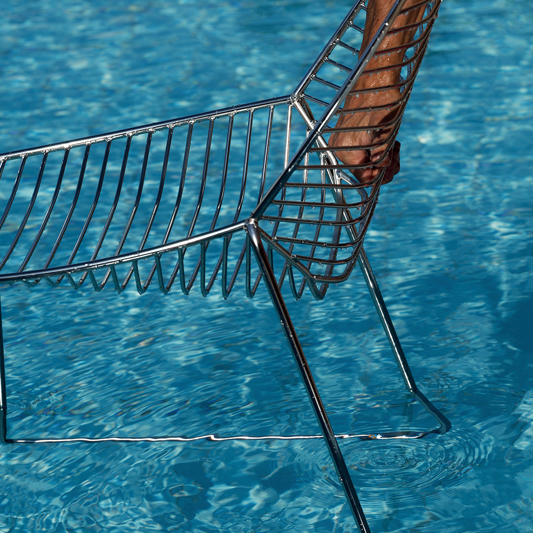 "Modern wire chair in shimmering silver against a vibrant blue water background."
