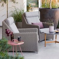 Gray woven patio chairs with light gray cushions, accented by a pink side table and vibrant green plants.