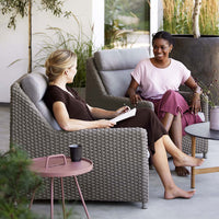 Two women relaxing in neutral-toned, woven chairs on a modern patio, surrounded by greenery and soft pastels.