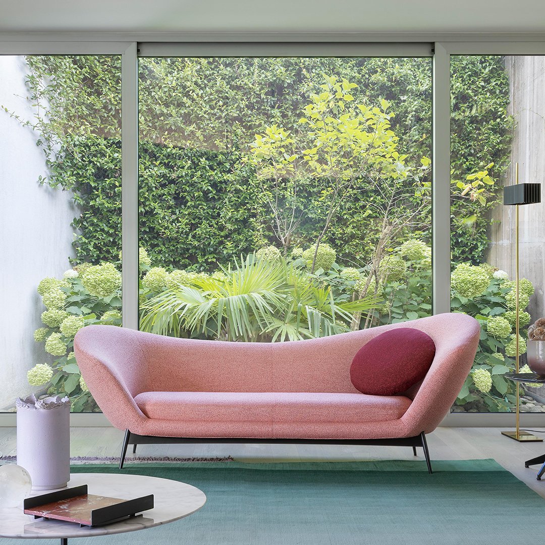 Pink and maroon modern sofa with curved design, set against lush greenery and a light green rug.