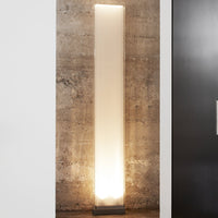 Sleek white floor lamp with a minimalist design, illuminated at the base, set against a textured concrete wall.