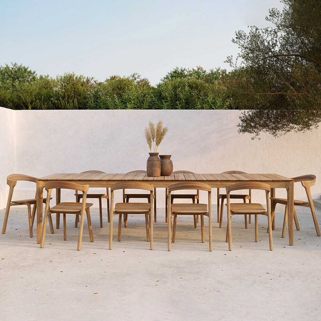 Natural wood dining table with light beige chairs, set against a soft white wall and green foliage backdrop.