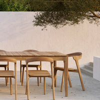 Natural wooden dining table and chairs with a smooth finish, set against a light backdrop and greenery.