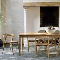 Natural wood dining table with light brown chairs, contrasting against a textured gray stone floor and soft white walls.