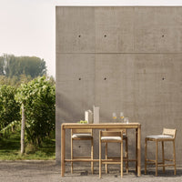 Natural wood furniture with light beige cushions against a textured grey concrete backdrop, set in a green vineyard.