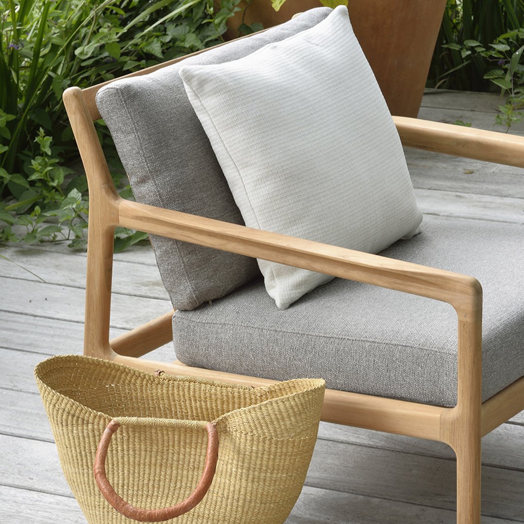 Gray and cream cushions on a light wood chair, complemented by a natural woven basket, set against greenery.