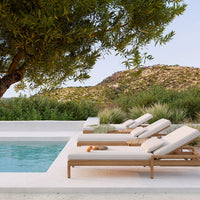 Beige lounge chairs beside a clear pool, surrounded by lush green foliage and a soft earth-toned landscape.