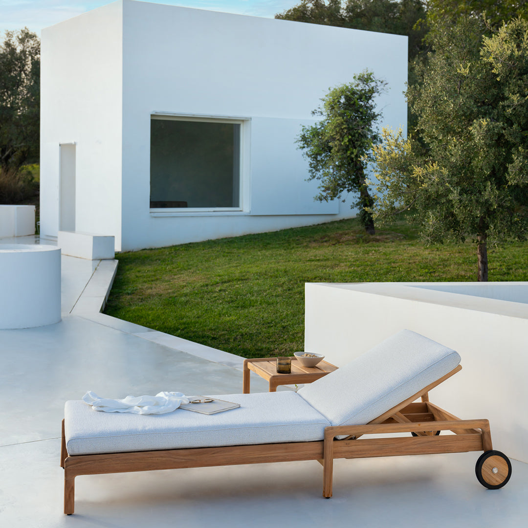 Light wood chaise lounge with white cushions against a modern white architectural backdrop and green landscape.
