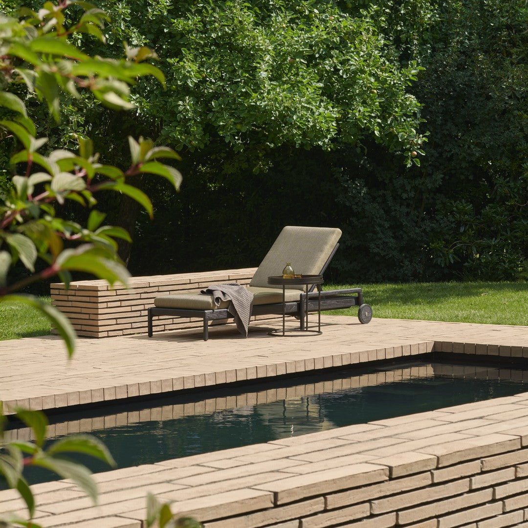 Neutral-toned lounge chair on a textured beige stone patio, surrounded by lush green foliage and a dark pool.