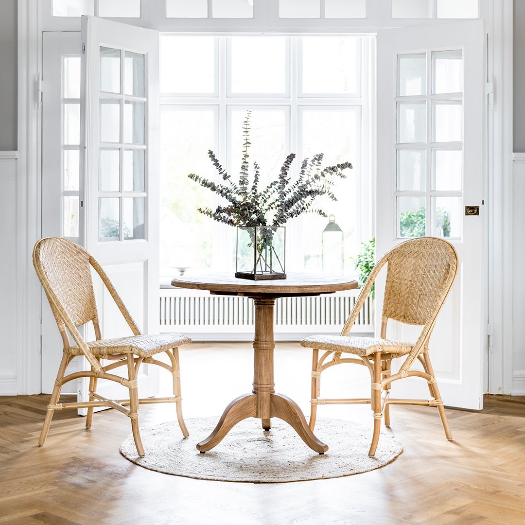 Light woven rattan chairs by a round wooden table, set against a bright, airy room with herringbone flooring.
