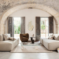 Neutral beige sofas with geometric patterns and a rich brown accent chair, set in a bright, airy interior with natural light.