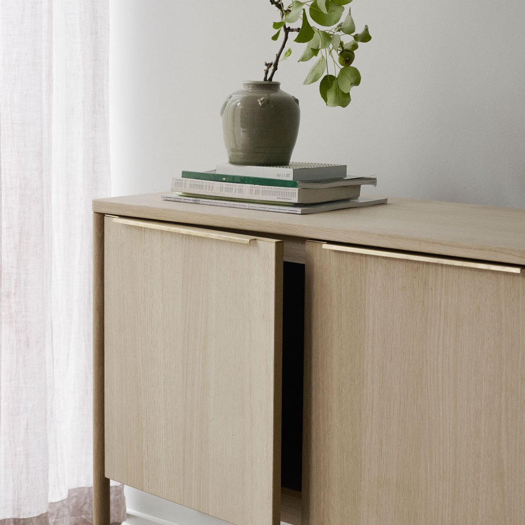 Light wood cabinet with natural grain, topped with a muted green vase and stacked neutral books, against a soft background.