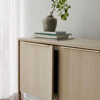 Light wood cabinet with natural grain, topped with a muted green vase and stacked neutral books, against a soft background.