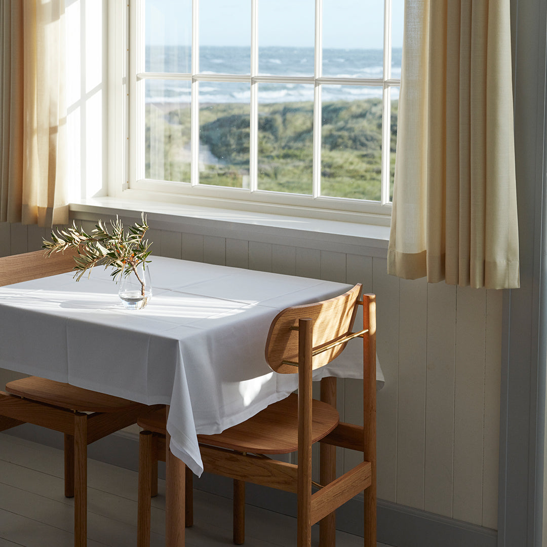 "Minimalist white tablecloth on a wooden table with pale yellow curtains, framed by a coastal view."