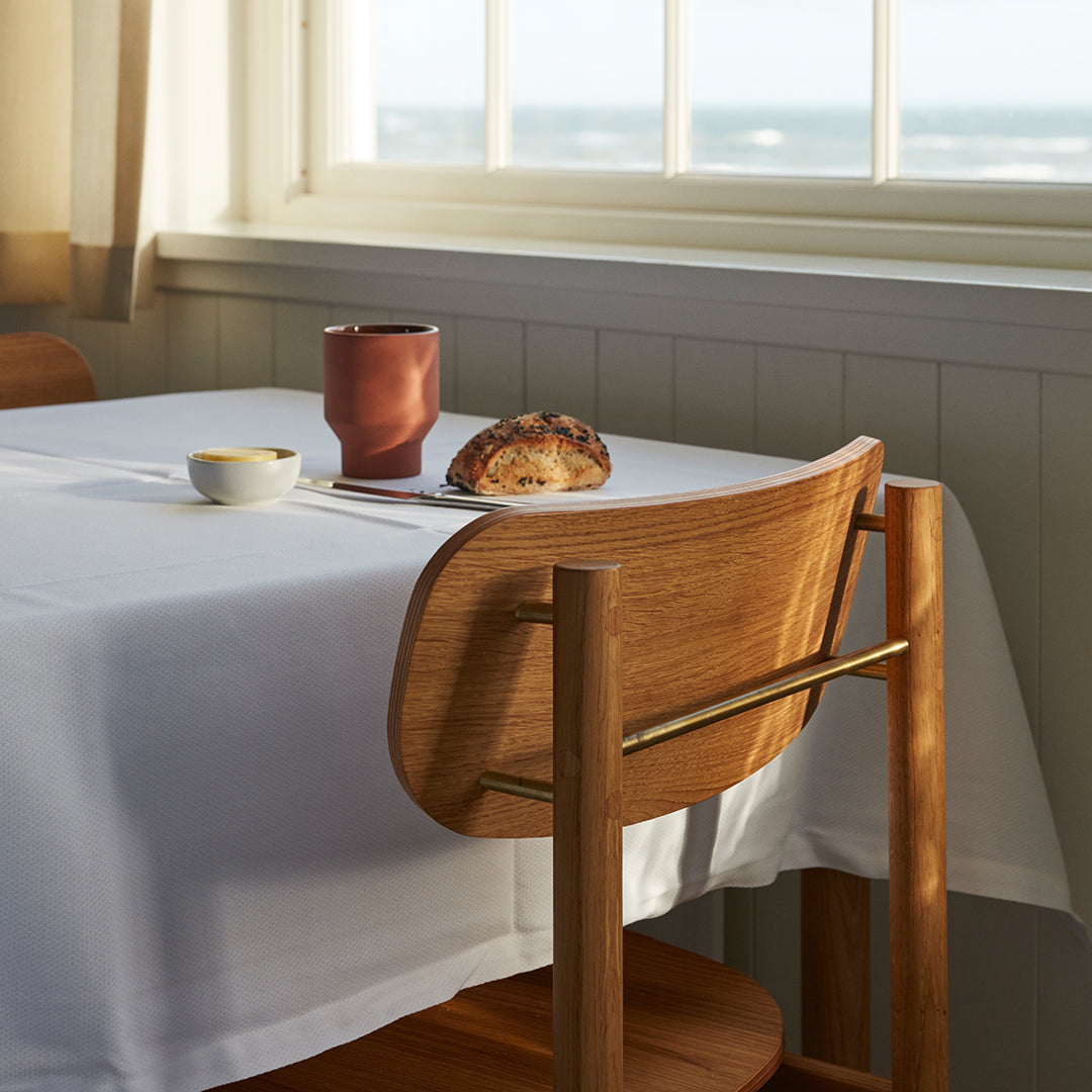 "Neutral white tablecloth with warm wood chair, soft brown croissant, and deep terracotta mug against a coastal backdrop."