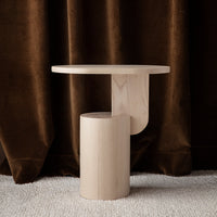 Light wood side table with a smooth round top and cylindrical base, against a rich brown velvet backdrop.