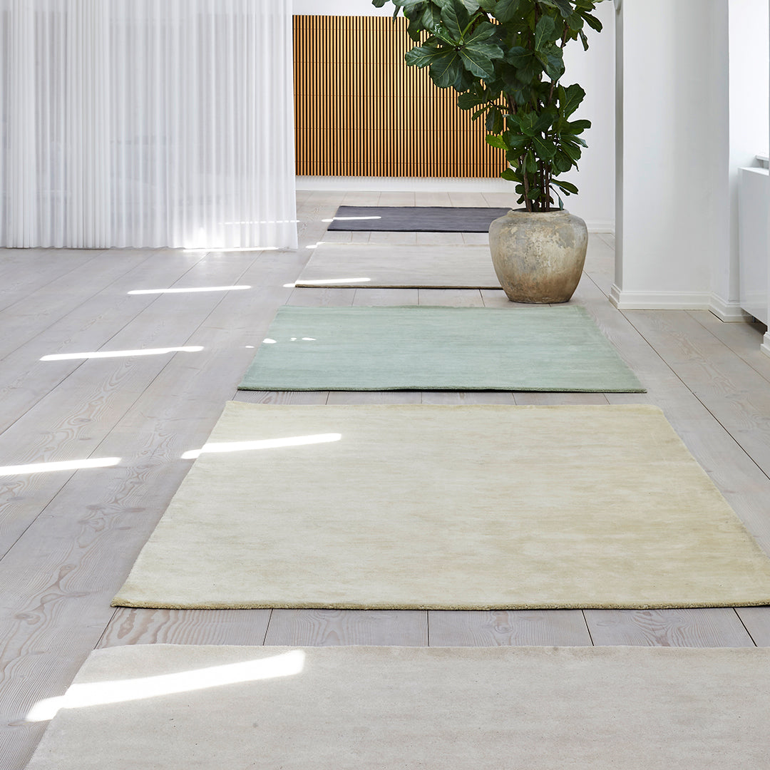 Assorted area rugs in soft green, cream, and gray tones, arranged on a light wood floor with natural light.