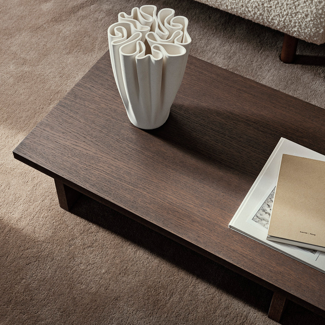 Dark wood coffee table with smooth surface, accompanied by a white textured vase and beige book on a soft carpet.