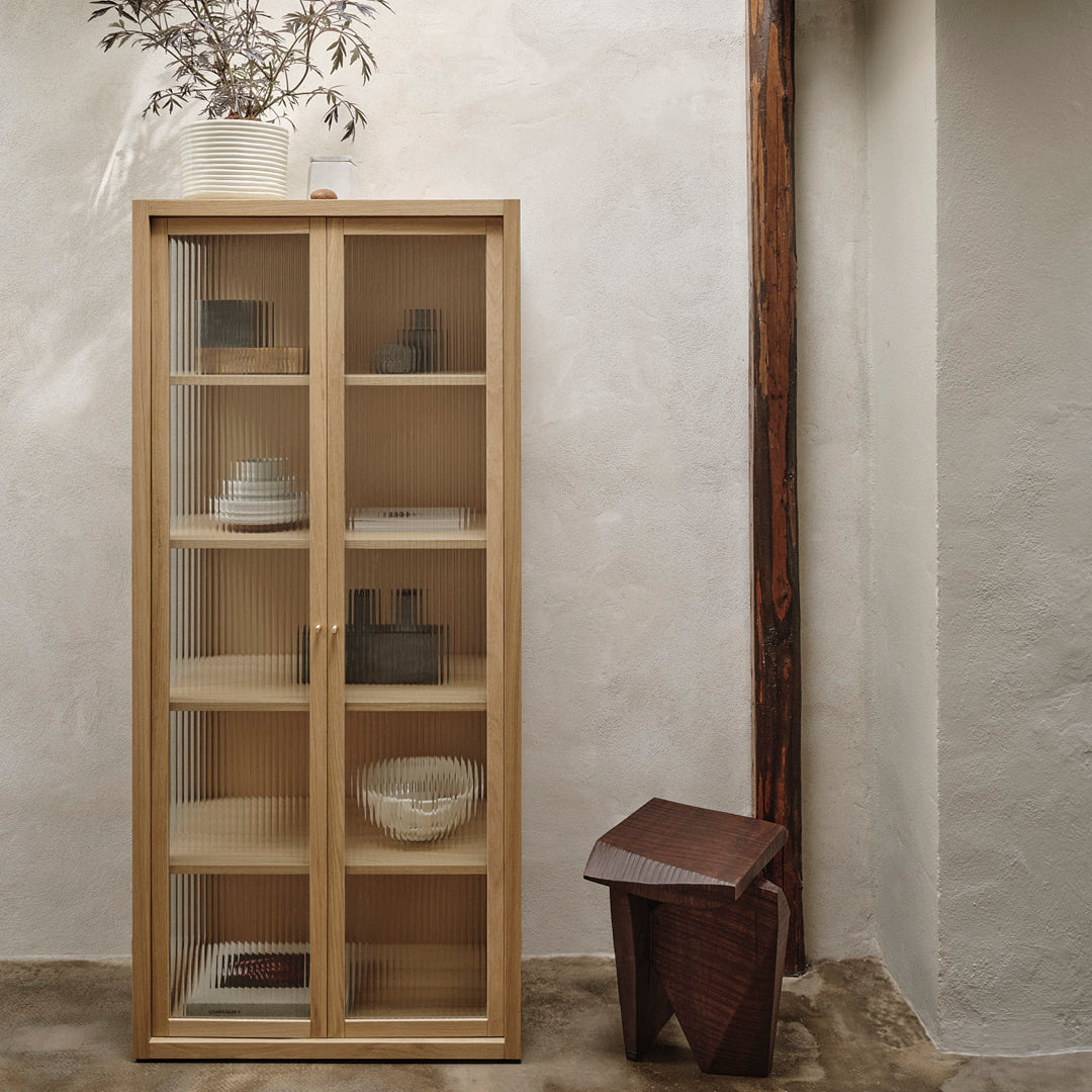 Light wood display cabinet with natural finish; clear glass doors showcase minimalist white and gray dishware.