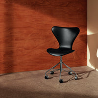 Black ergonomic chair on chrome wheels against a warm wood panel background and neutral carpet. Modern design.