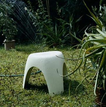 White plastic garden stool in a lush green setting, with water droplets on its surface and surrounding foliage.