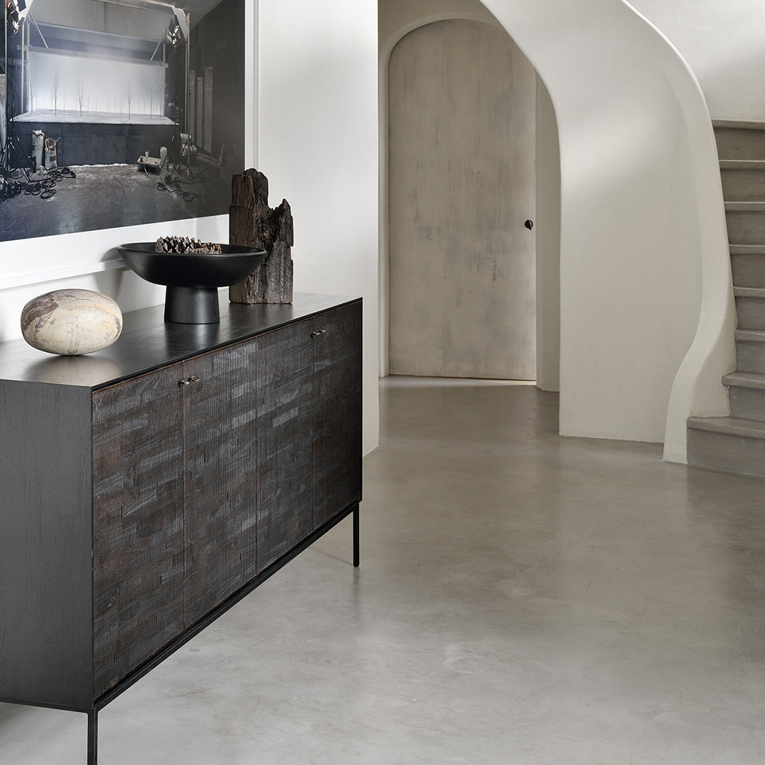 Black woven cabinet on gray concrete floor with minimalistic white wall and curved staircase. Modern and sleek design.
