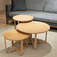 Set of three round wooden nesting tables in light oak finish with natural grain pattern, complementing a gray sofa.