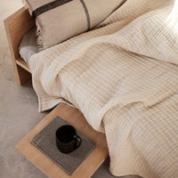 Beige textured blanket with a subtle grid pattern, paired with a neutral pillow and wooden bedside tray.