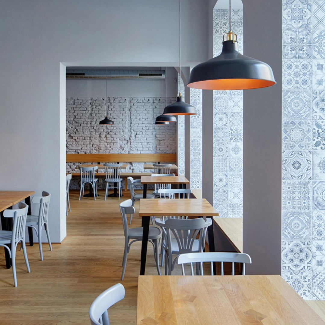 Modern cafe interior with white patterned wallpaper, light wood tables, and gray metal chairs, accented by black pendant lights.
