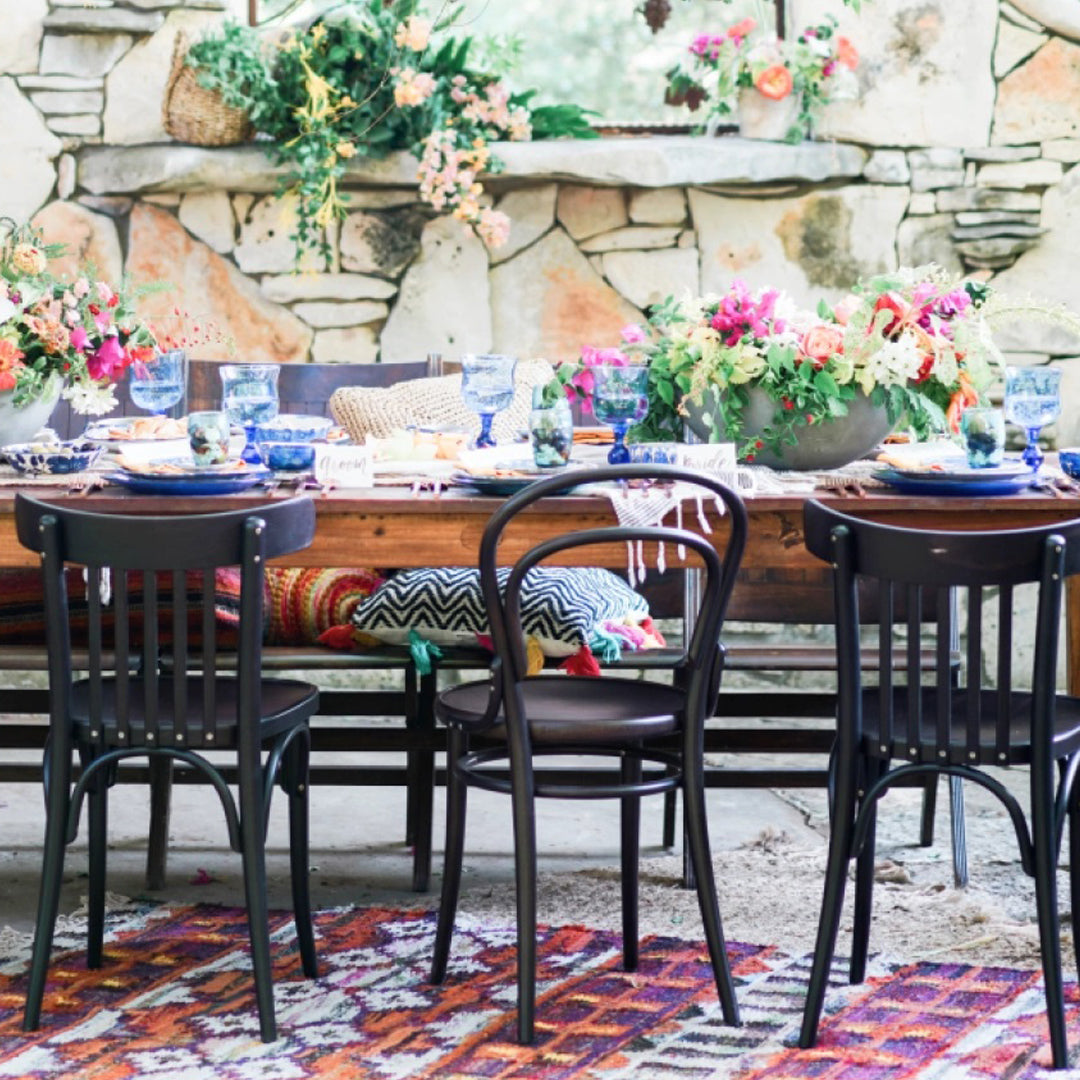 "Wooden table set with blue tableware, vibrant floral patterns, and a colorful geometric rug in an outdoor setting."