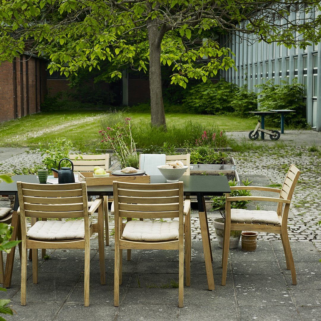 Wooden outdoor dining set featuring beige cushions and a sleek black table surrounded by green foliage.