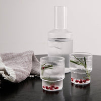 Clear ribbed glass water carafe and tumblers with fresh herbs and berries, set on a dark wooden table with a textured towel.