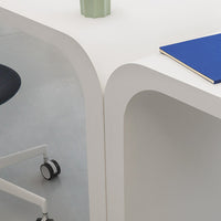 White desk with rounded edges, a blue notebook, and a green vase, featuring a minimalist design on a gray floor.