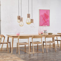 "Light wood dining table and chairs with pink and amber pendant lights against a textured white wall."