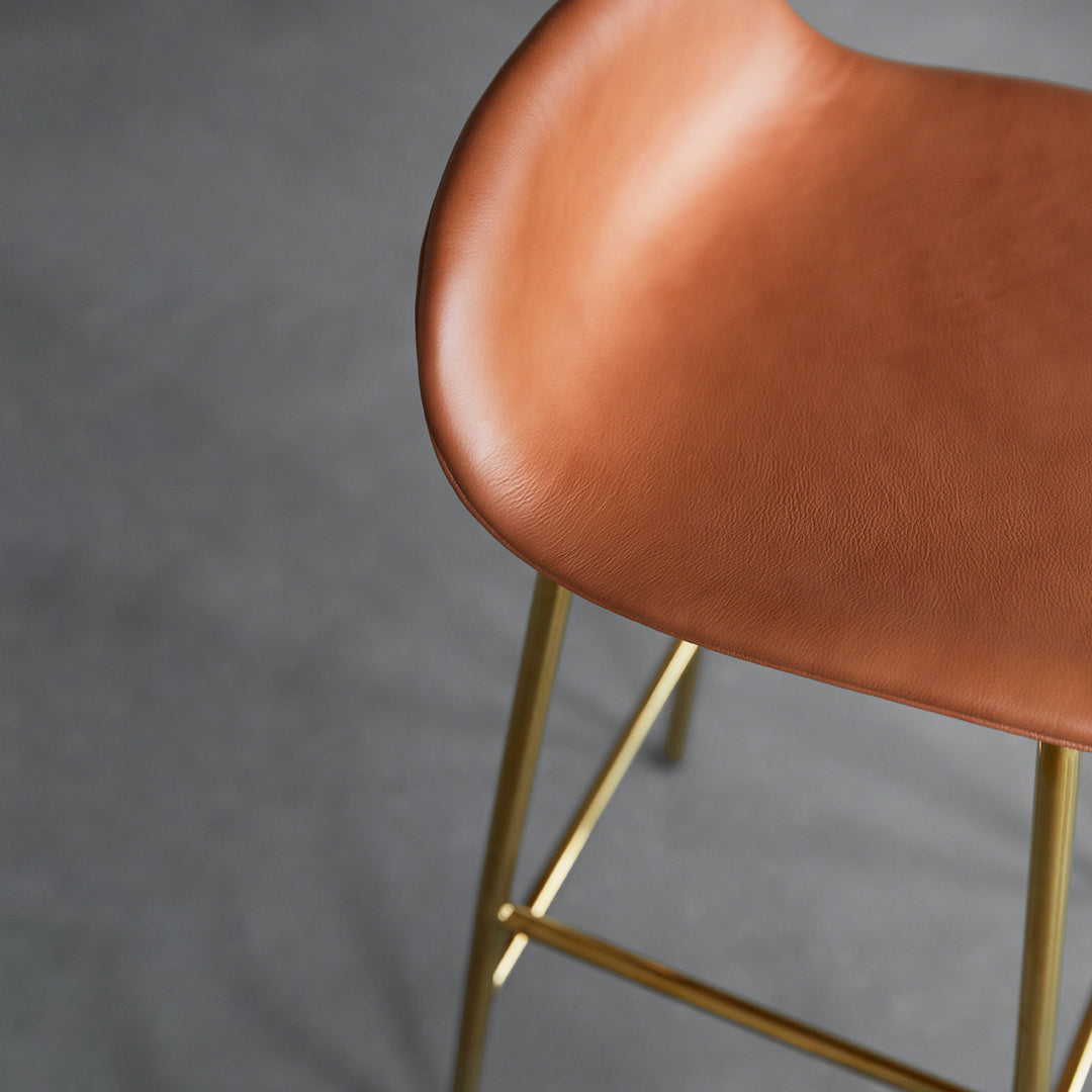 Brown leather bar stool seat with smooth surface and stylish gold metal legs on a gray background.