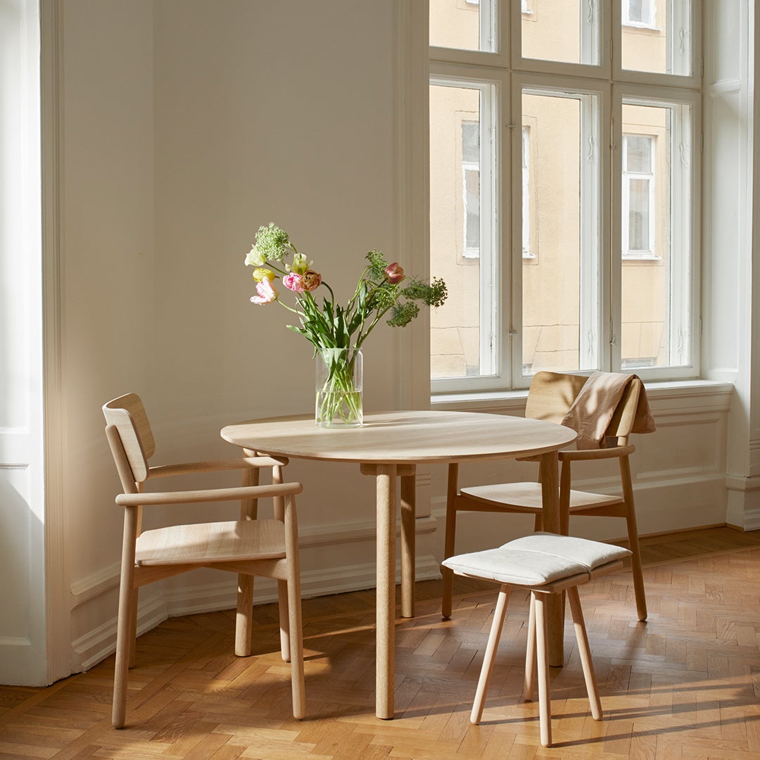 Light wooden dining table surrounded by beige chairs and small white stool, with a flower vase adding a pop of color.