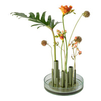 Colorful floral arrangement with orange blooms, green leaves, and a clear glass base featuring green tubes.