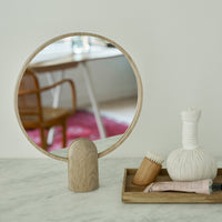 Wooden round mirror with a light wood base, reflecting soft beige and white decor, featuring subtle pink patterns in background.